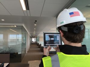 A person wearing a safety helmet and reflective vest using a tablet to implement technology in an office corridor.