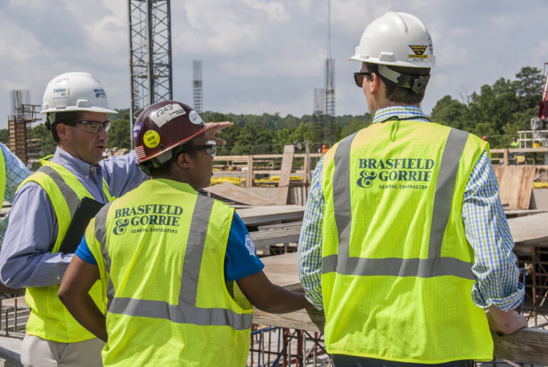 Three construction workers in hard hats and high-visibility vests are discussing on a construction site.