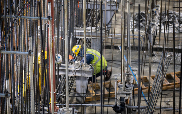 A construction worker wearing a hard hat and high-visibility jacket is engaged in work amidst scaffolding and rebar.