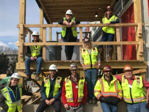 A group of construction workers in safety gear, including women in construction, posing on a wooden structure at a construction site.