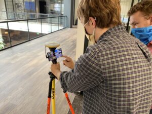 Two individuals operating a construction technology surveying instrument indoors.