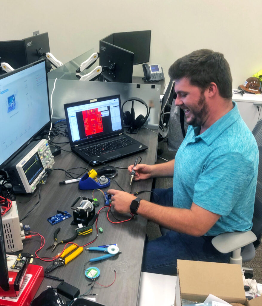 A man smiling while working on electronic equipment in an office setting with various tools and computers on the desk, embodying the spirit of tech industry leaders.