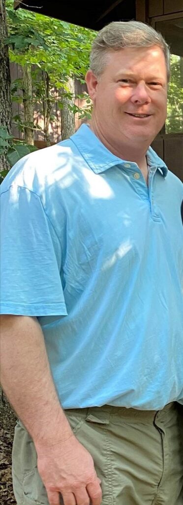 Man in a blue shirt smiling outdoors at a challenging construction site.