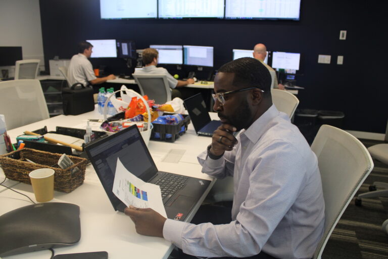 A man in a striped shirt reviews a document at his desk in a busy office environment, illustrating a mentorship success story.