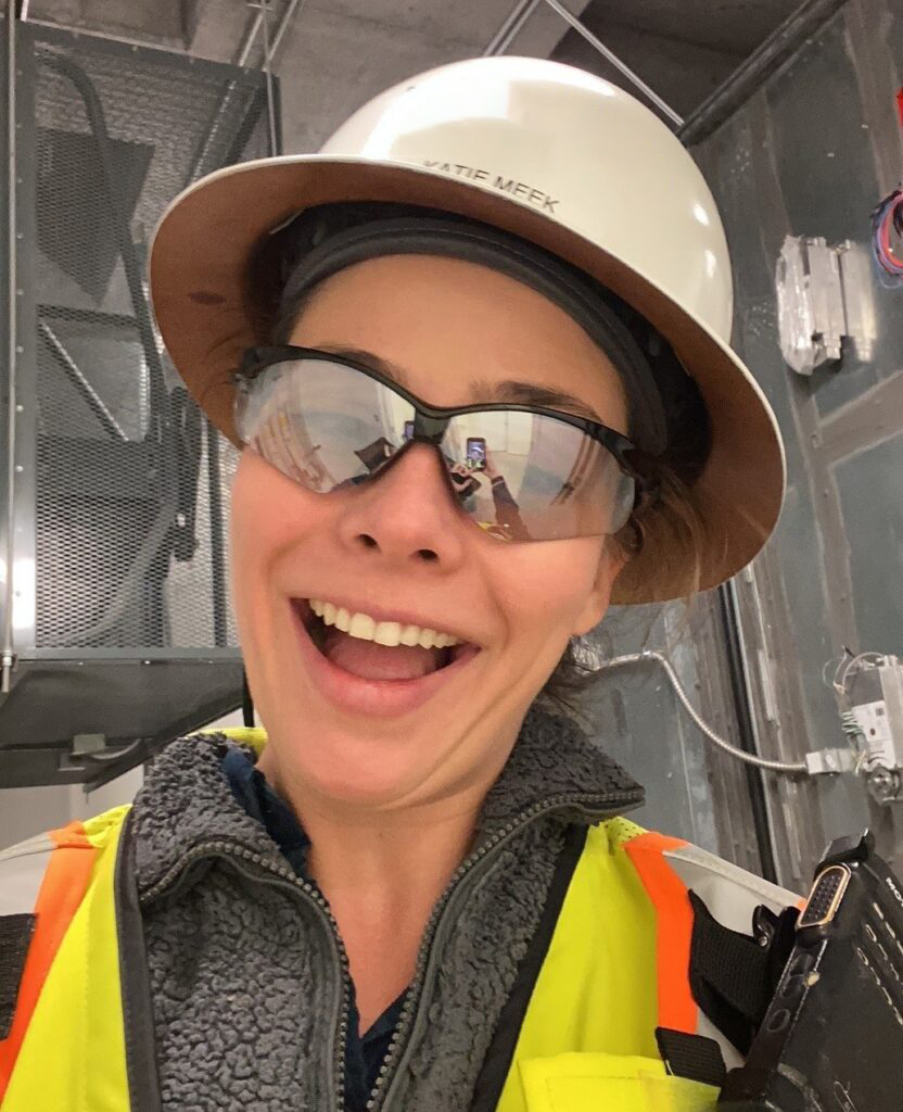 A smiling woman wearing a hard hat, safety glasses, and a high-visibility vest on a construction site during Construction Week.