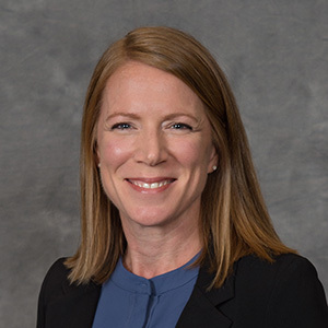 A professional headshot of a smiling woman with long hair, wearing a blue top and a blazer, symbolizing progress during Women in Construction Week.