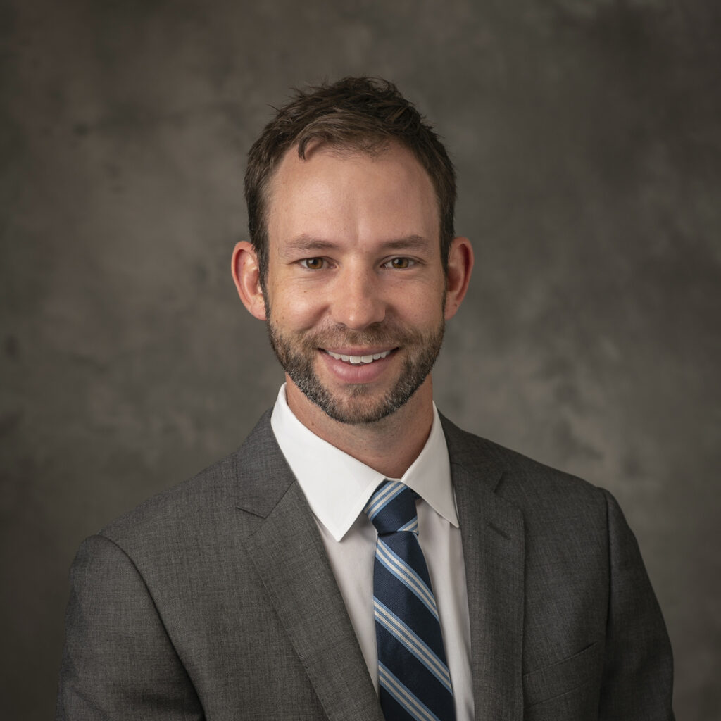 A professional portrait of Jason Weeks, a smiling man in a suit and tie against a textured gray background.