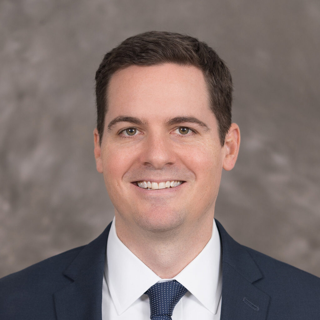 A man in a business suit with a tie smiling at the camera against a grey background, symbolizing success in the Science and Technology Market Sector.