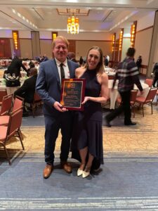 A man and a woman dressed in formal attire, smiling and holding a "Contractor of the Year" award at an indoor event.