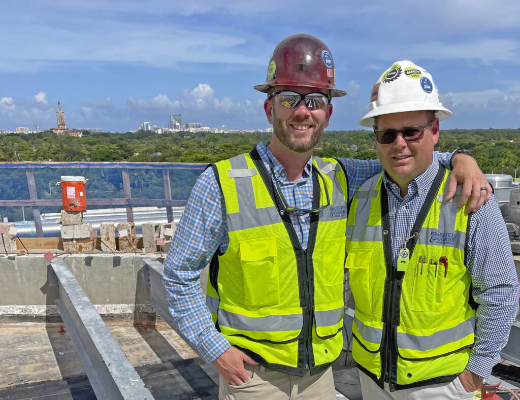 Two construction workers in hard hats and reflective vests posing for a photo with a South Florida construction site in the background.