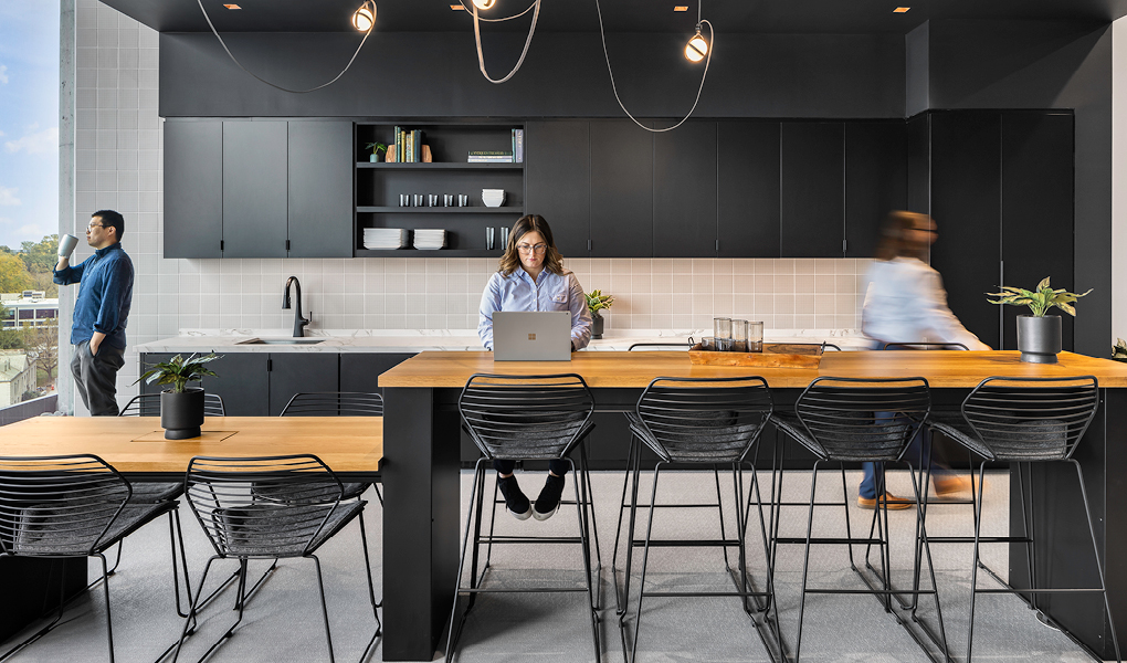 Three individuals in a modern kitchen within Midtown Union, Atlanta, with one seated at a central island working on a laptop, another standing by the counter, and the third person in motion.