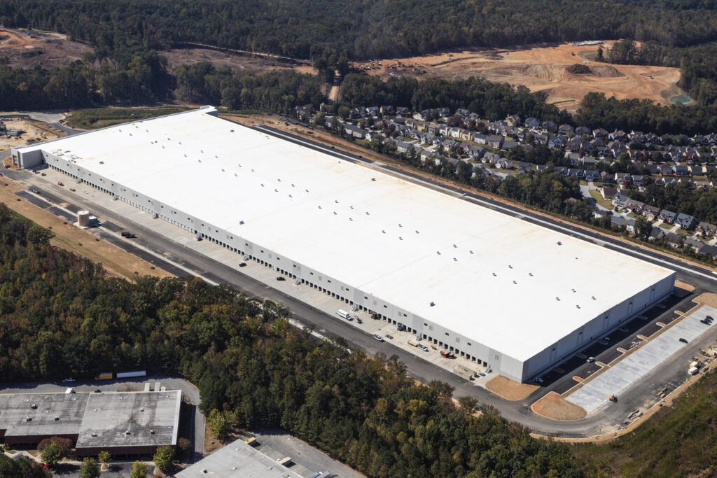 Aerial view of a large distribution center under construction with surrounding parking and a residential area in the background.