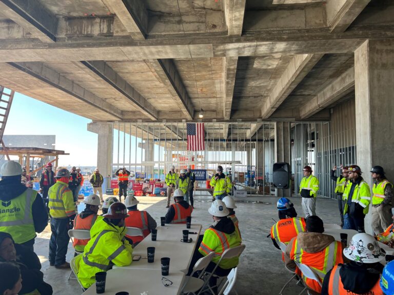 Construction workers gathered for a meeting under an unfinished concrete structure at a Vondace Contracting manufacturing facility, celebrating a construction milestone with an American flag hanging in the background.