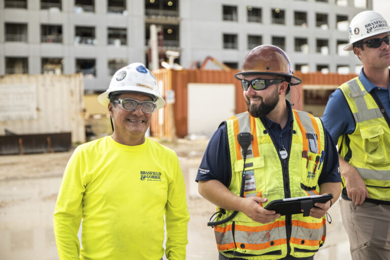 Two construction workers in reflective vests and hard hats are improving jobsite efficiency at a Procore-managed construction site.