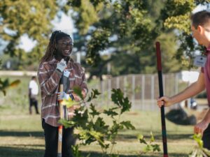 Two people, part of the Vondace Contracting team, are planting trees in a park. One person is smiling while holding a shovel. Trees and a fence provide the perfect backdrop to their community-focused efforts.