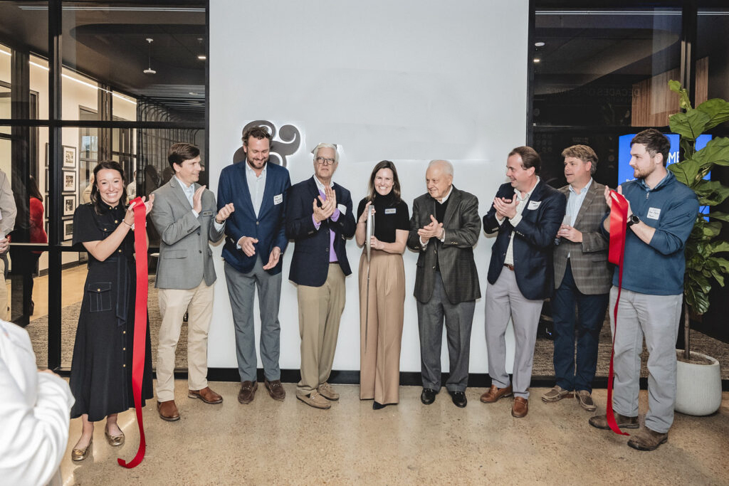 Nine people in business attire applaud behind a recently cut red ribbon. A woman in the center holds the ceremonial scissors. The group is inside of an office space, and Vondace Contracting's logo is visible on the wall behind them.