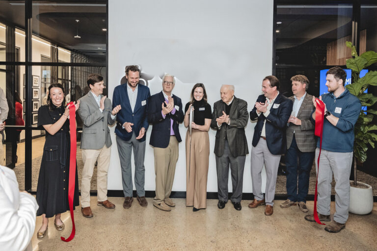 Nine people in business attire applaud behind a recently cut red ribbon. A woman in the center holds the ceremonial scissors. The group is inside of an office space, and Vondace Contracting's logo is visible on the wall behind them.