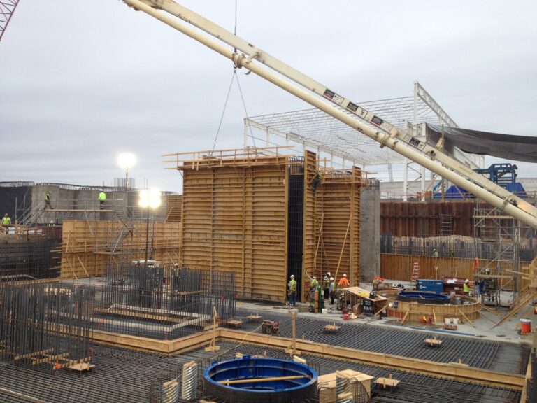 Construction site with workers and equipment near a large concrete formwork under a crane, with early evening lighting in the industrial sector.