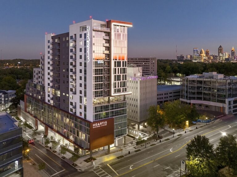 Modern multi-story senior living building at twilight with illuminated windows and surrounding cityscape in the distance.