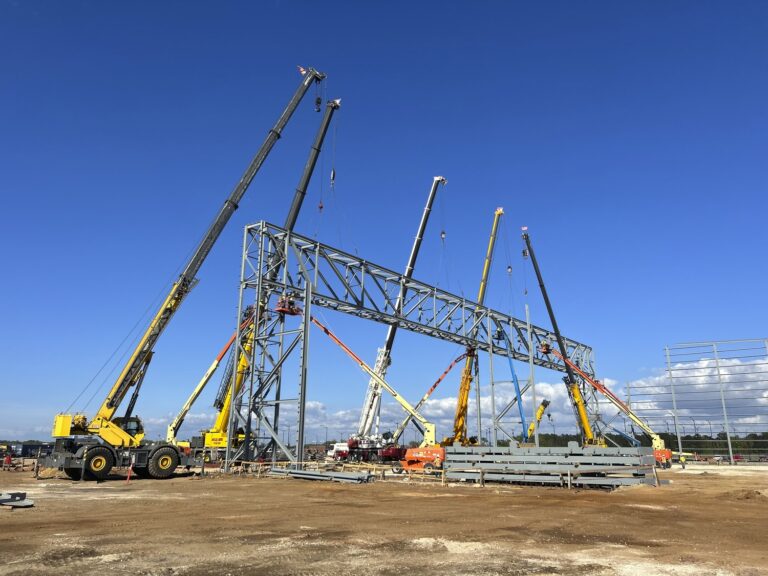 Construction cranes erecting a large steel structure for a heavy/civil construction supersector project on a clear day.