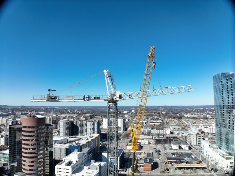 Two cranes, part of a Nashville project by Brasfield and Gorrie, are diligently at work on a high-rise construction site, set against a clear blue sky, with urban buildings and distant landscapes completing the scene.