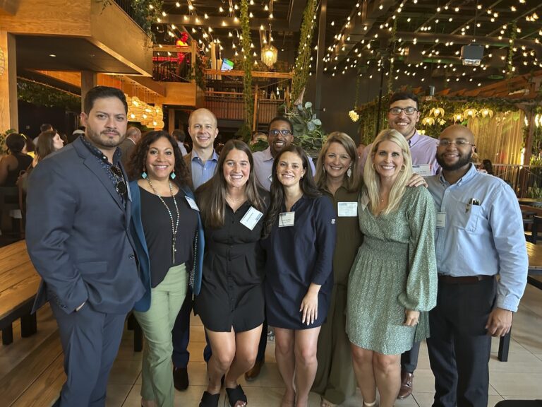Group of smiling people posing for a photo at a social event with string lights in the background.