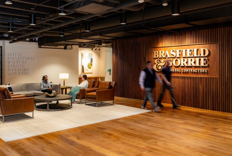 Two people seated and conversing in a modern office lounge with a blurred figure walking by a company branded wall.