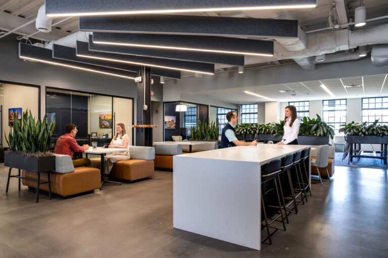 Modern Greenville Office Brasfield and Gorrie space with people talking and working; features large windows, plants, and contemporary furnishings, including a long table with chairs and lounge seating.