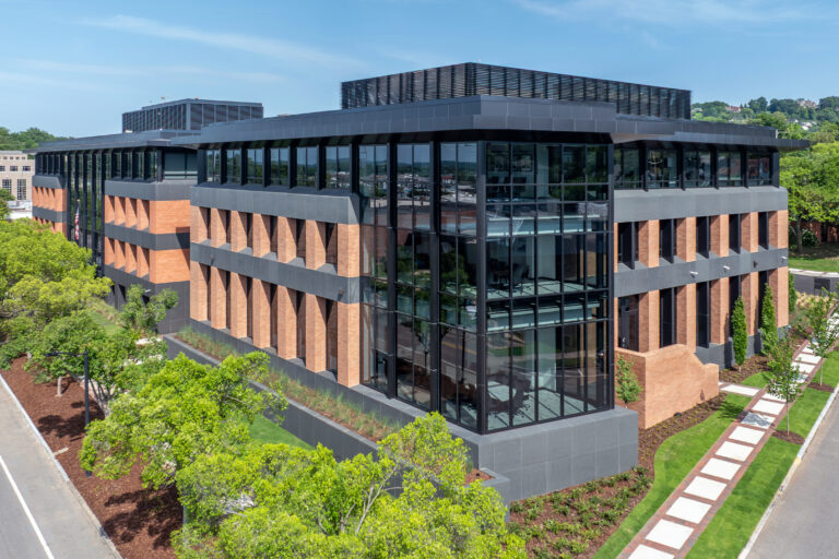 Modern office headquarters with large glass windows and brick accents, surrounded by trees and landscaped pathways on a sunny day.