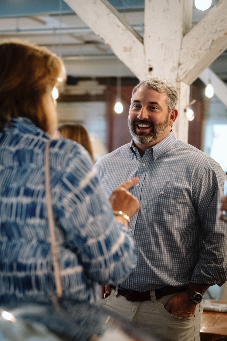 A smiling man in a checkered shirt converses with a person whose back is to the camera in a room with wooden beams.