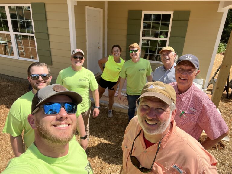 Group of smiling volunteers in matching green shirts taking a selfie at a construction site.