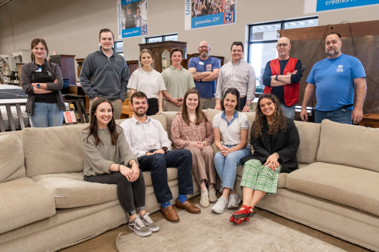 In a Nashville office, a group of people, some seated on a large beige sofa and others standing behind, pose for a photo. The setting is bright and welcoming, with tables and cabinets visible in the background.