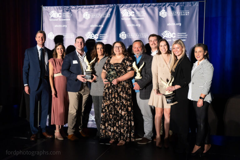 A group of well-dressed individuals from the Nashville office stands together, proudly holding awards, in front of a backdrop adorned with logos and text.