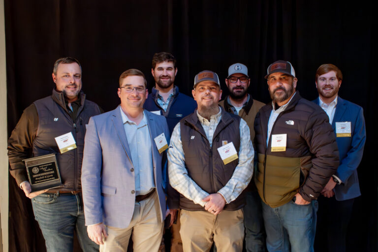 Seven men standing together, some with name badges, dressed in casual and business attire against a dark backdrop. Among them, one man holds a plaque.