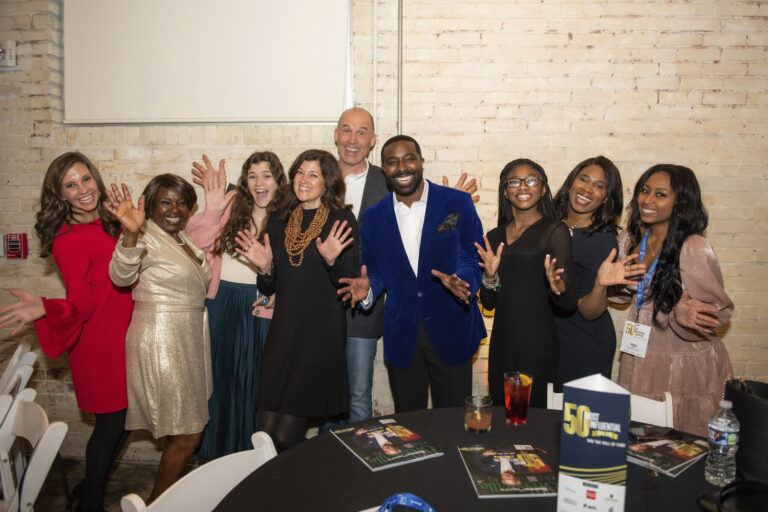 Group of eight people posing with smiling faces and open hands, as if to show excitement or celebration, at a social event with a brick wall background.