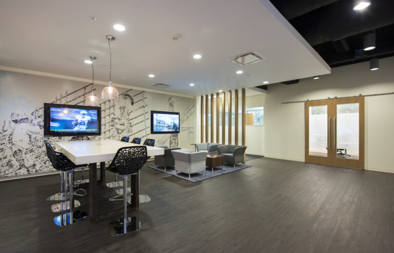 Modern office lounge area featuring a high table with stools, comfortable seating, mounted televisions, and decorative lighting.
