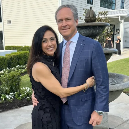 Keith Johnson stands in a blue suit beside a woman in a sleeveless black dress, both smiling as they pose in front of a fountain outside a building.