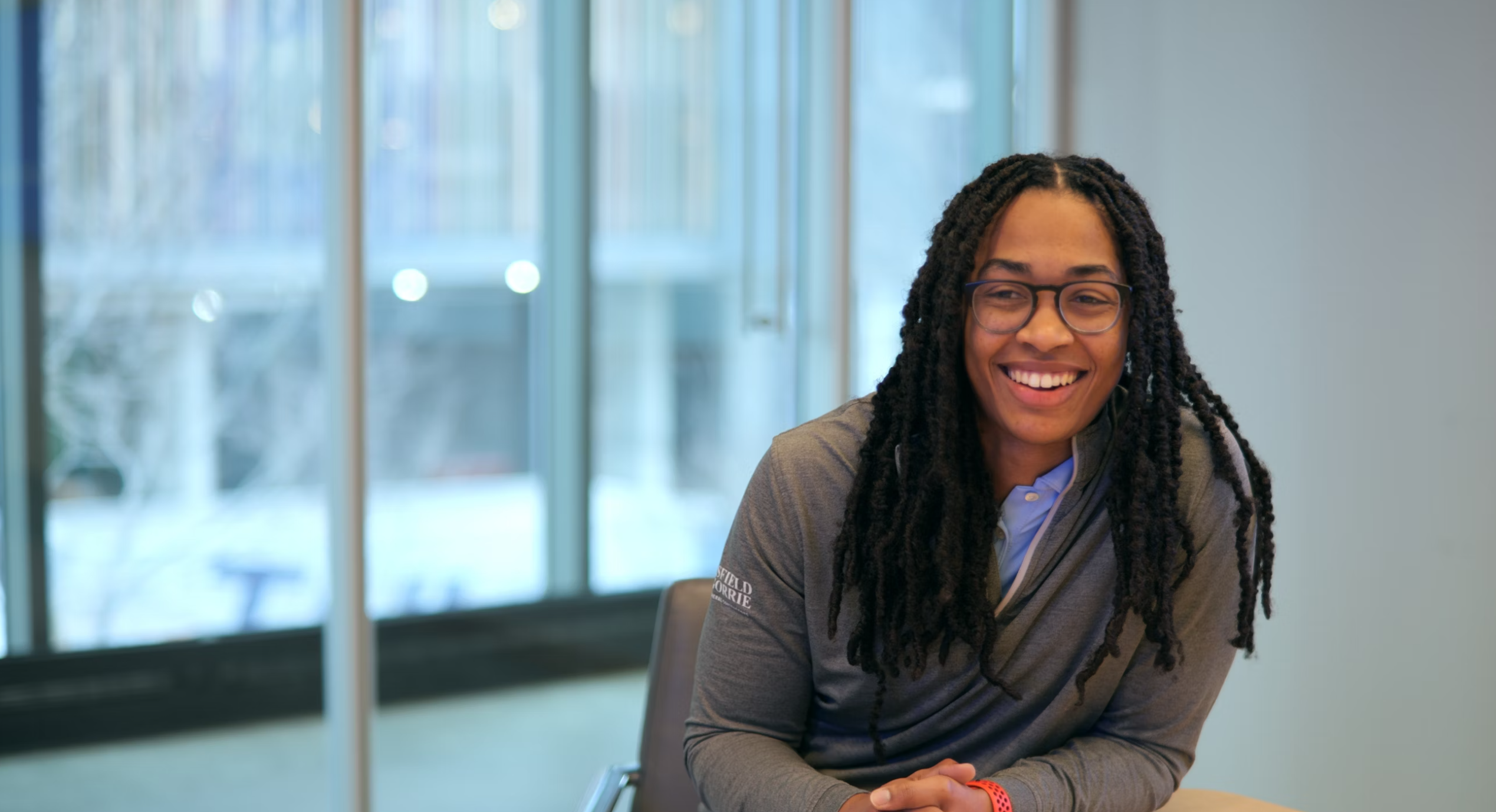 A person with braided hair smiling at the camera in a room with window views of a snowy outside, considering careers.