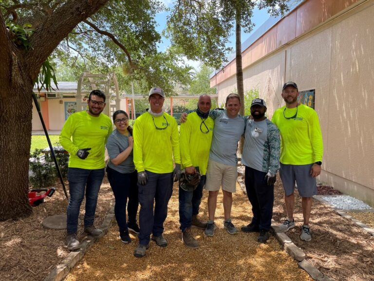 A group of eight adults in bright green shirts posing together outdoors, likely volunteers or workers from the same organization, with trees and a building in the background.