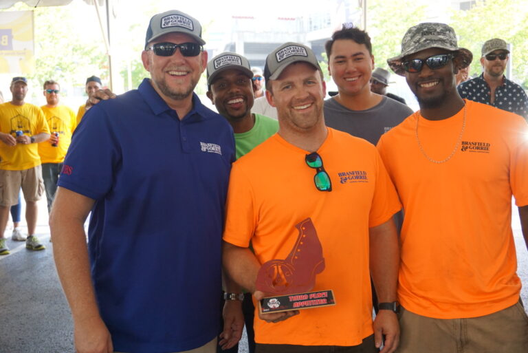 A group of men smiling outdoors, with one holding an award trophy.