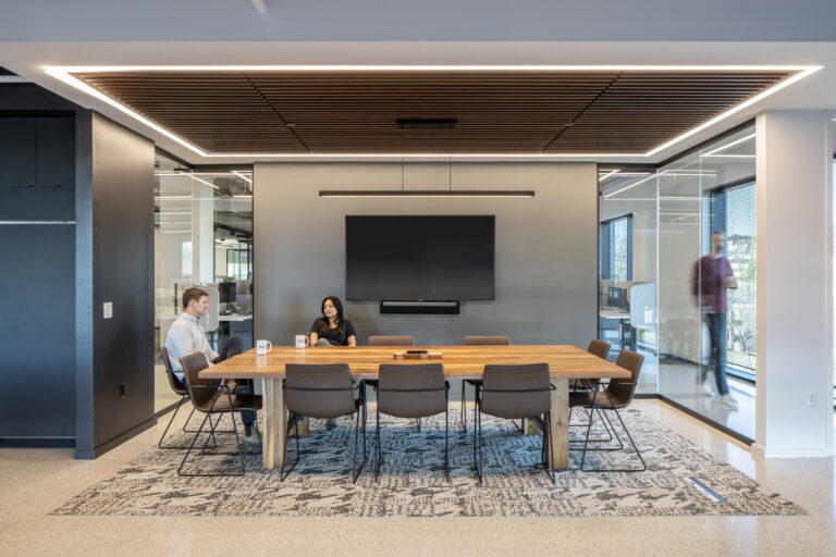 Modern office meeting room with two individuals seated at a conference table, a wall-mounted screen, and a person walking by in the background.