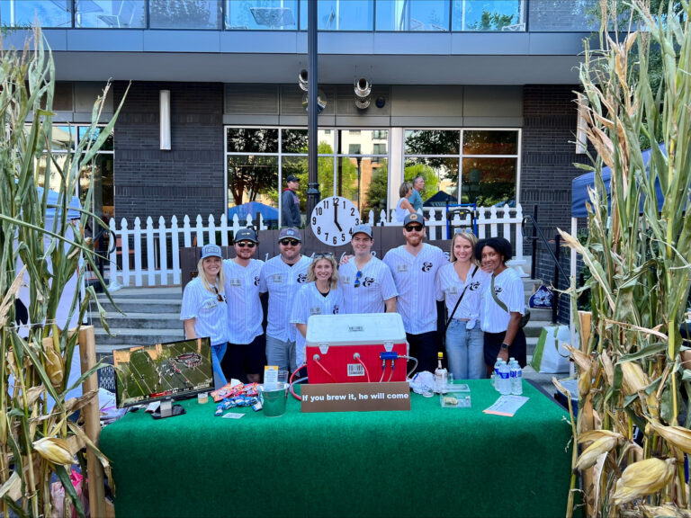 A group of people at an outdoor event stand behind a booth with promotional materials, beverages, and cornstalks.