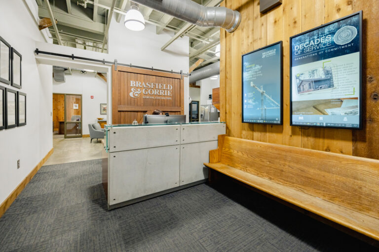 Modern office lobby with a reception desk, wooden bench, and informational digital display.