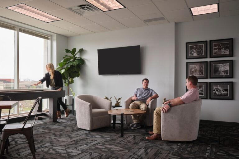 Three colleagues in a modern office lounge in Jackson, Mississippi, one looking out the window towards local attractions, two sitting and having a conversation.