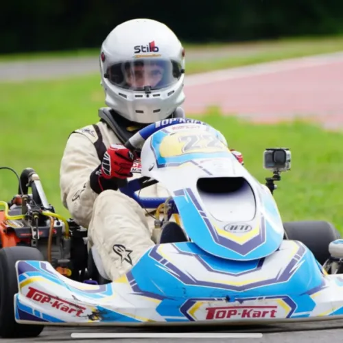 Person in a white racing suit and helmet driving a blue and white go-kart with “TOP-Kart” branding on a race track near grass.