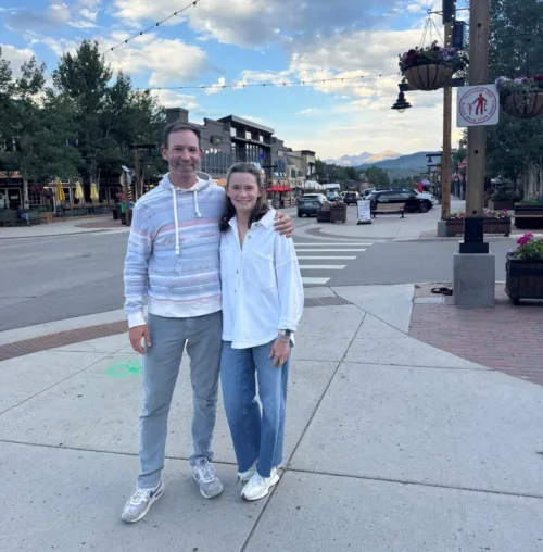 Two people stand smiling on a street corner in a small town with shops, trees, and mountains in the background under a partly cloudy sky.