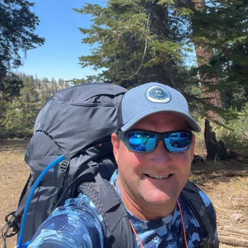 Man wearing a blue cap and sunglasses takes a selfie while hiking in a forested area with tall trees and a clear blue sky.