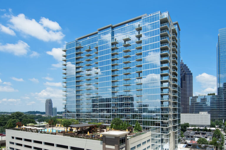 Modern glass high-rise residential building in an urban setting with clear blue sky and surrounding greenery