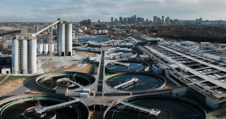Aerial view of an urban wastewater treatment plant with multiple circular settling tanks, concrete structures, and industrial buildings, all expertly constructed by Vondace Contracting, with a city skyline visible in the background.