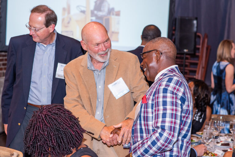 Two gentlemen shaking hands at a formal event, encircled by other participants and a seated dining area in the background.
